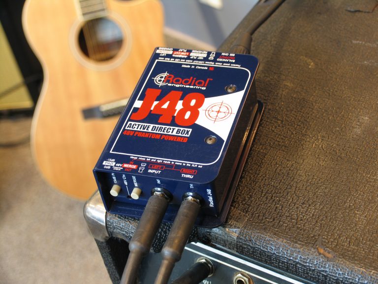 A J48 direct box sitting on a guitar amplifier beside an acoustic guitar